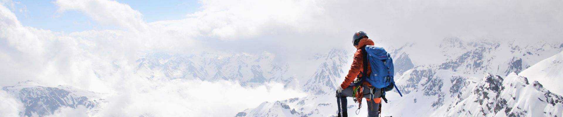 Ein Bergsteiger steht in voller Montur auf dem Berggipfel und schaut über die verschneiten Wipfel.